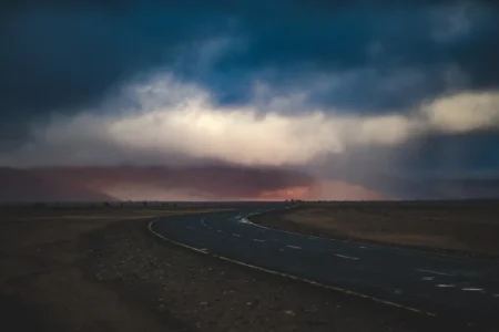 A winding road through a desert landscape under a dramatic, stormy sky with clouds gathering.