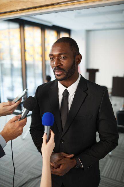 Businessman in suit speaking to journalists with microphones indoors.