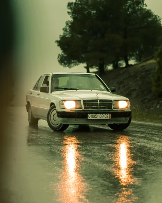 A classic white Mercedes Benz driving on a wet road in Algeria, reflecting in the rain-soaked surface.