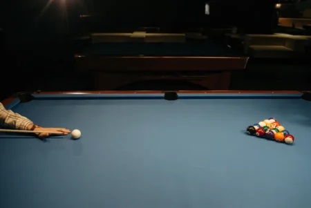 A player's hand aiming at a cue ball on a billiard table with racked balls.