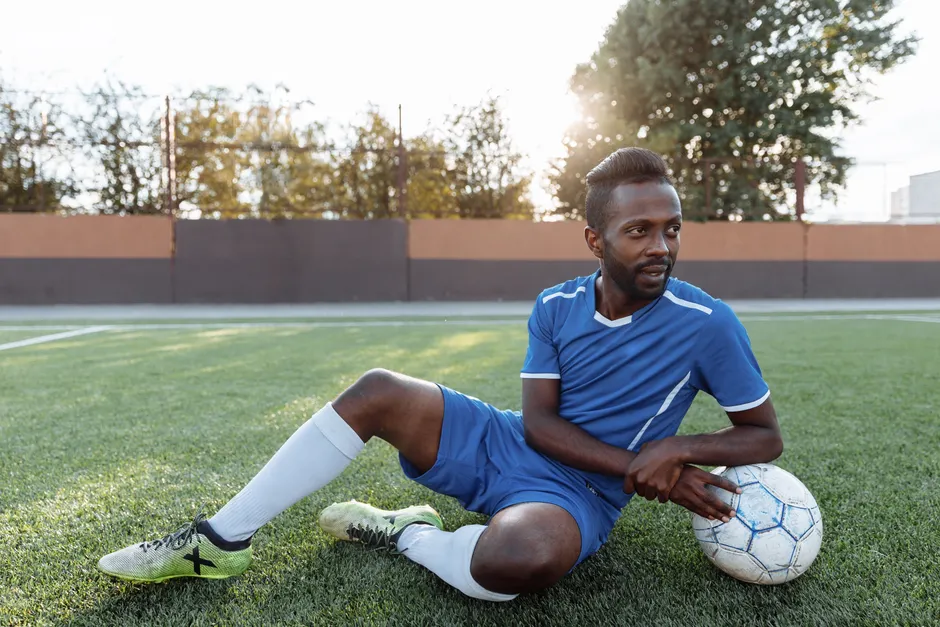 Blessure de Mbappé au genou : Madrid attend A soccer player in blue jersey sitting on a green field holding a ball, enjoying a sunny day.