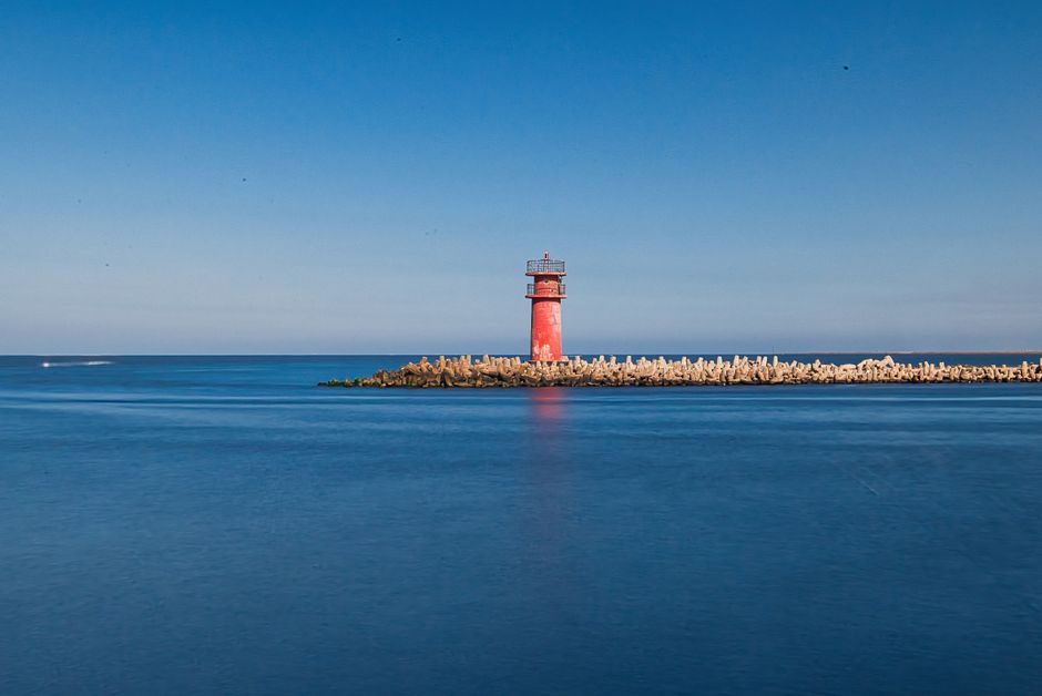 A vibrant red lighthouse stands on a tranquil coastline in Ras El-Bar, Egypt, under a clear blue sky.