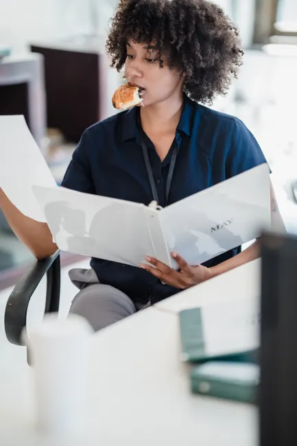 A woman multitasks by eating a snack while reviewing documents in a busy office setting.