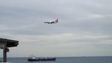Une erreur d’IA militaire a failli provoquer une interception Airplane approaching over the sea with a cargo ship below under a cloudy sky.
