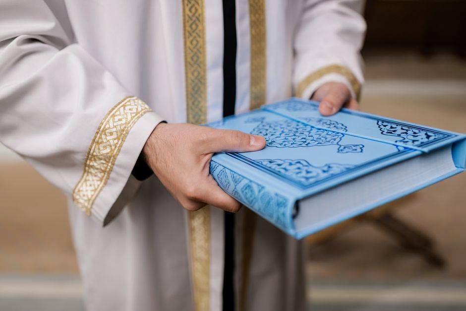 Tarawih à l’étranger : 539 candidats évalués en Algérie Close-up of a man holding a blue Quran, dressed in a traditional robe, signifying Muslim culture and religion.