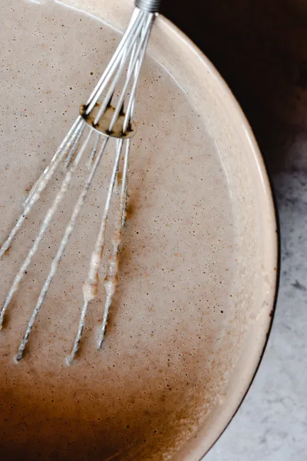 Close-up of a whisk mixing pancake batter in a ceramic bowl on a kitchen counter.