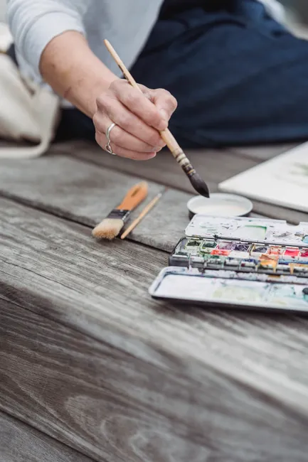 Close-up of an artist's hand using watercolors on a wooden bench outdoors. Artistic and creative vibe.