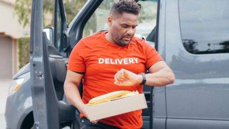 Suivi UPS : lire un statut, vérifier un retard et agir Delivery driver checks watch while carrying package from van outdoors.