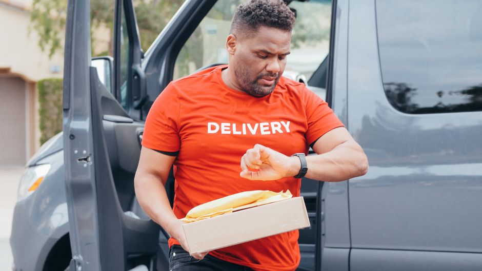 Suivi UPS : lire un statut, vérifier un retard et agir Delivery driver checks watch while carrying package from van outdoors.