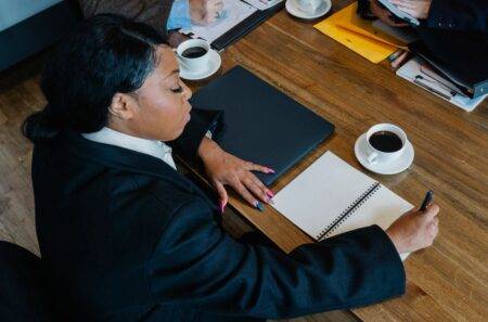 From above of black woman in formal clothes sitting at table during business meeting and writing notes in notebook