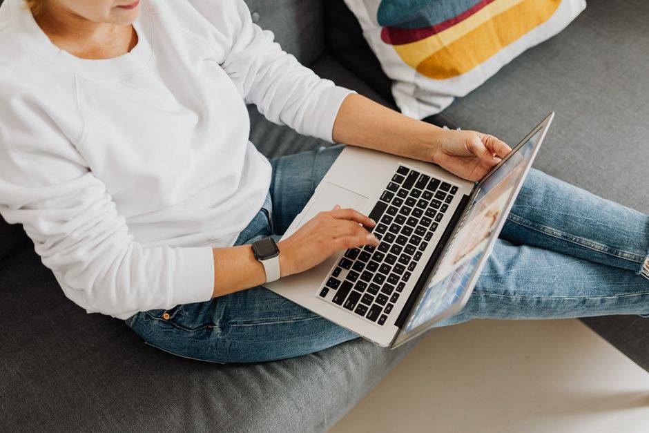 Hadj 2027 : l’Algérie appelle à ouvrir un compte numérique Person in casual attire using laptop on cozy grey sofa, creating a home workspace vibe.