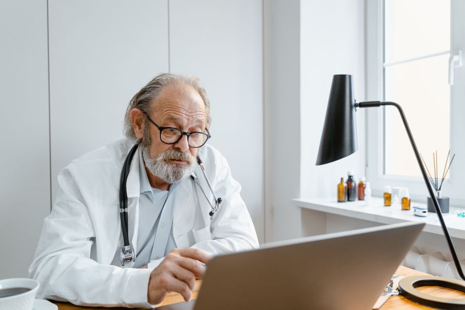 Carmen Aprea à Angri : téléconsultation et paiement Senior male doctor in lab coat using laptop for video call in clinic office.