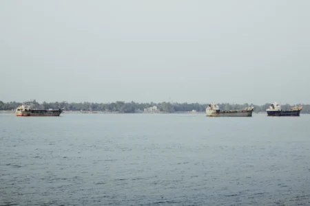 Three cargo ships float peacefully on a serene river under a clear sky, with a lush forested shoreline.