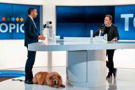 El Bilad, entraide, confidences et football algérien Two hosts discussing at a modern TV studio with a dog laying by. Engaging professional setup.