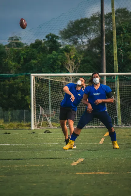 Two men are playing football outdoors on a field wearing face masks for safety measures. Action-oriented shot.