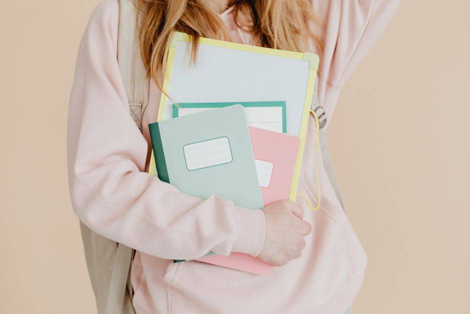 Young student in pink hoodie holding colorful notebooks, ready for school.